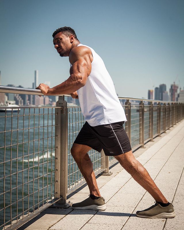 Larry Wheels standing against a grilled fence in a white tank top, black shorts, and sneakers.